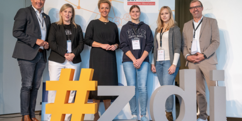 District Administrator Frank Rock, Mayor Susanna Stupp, Minister Ina Brandes, student Lena Engel, chemical laboratory assistant Pia Münstermann and zdi coordinator Axel Tillmanns stand next to each other behind the #zdi logo