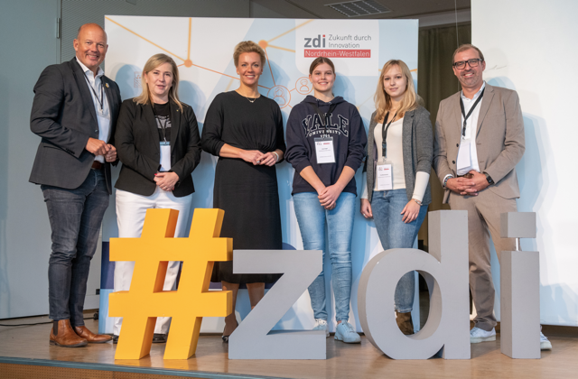 District Administrator Frank Rock, Mayor Susanna Stupp, Minister Ina Brandes, student Lena Engel, chemical laboratory assistant Pia Münstermann and zdi coordinator Axel Tillmanns stand next to each other behind the #zdi logo