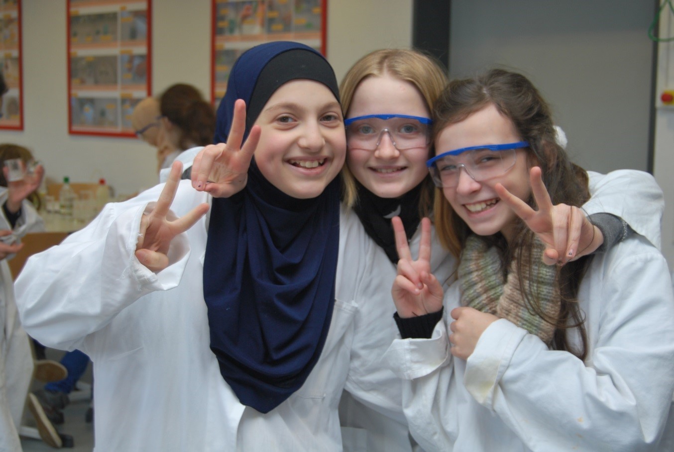 Три девушки в лабораторных халатах показывают знак победы.