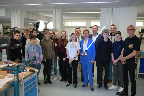 The photo shows a group of people, including many students. Minister Ina Brandes is in the front center. She is holding a blue glowing lightsaber.