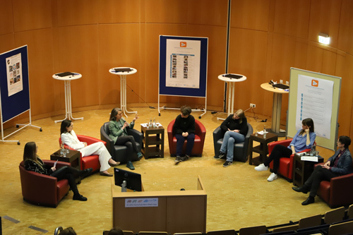 The photo shows all seven participants in the panel discussion sitting in armchairs in a semicircle. Kerstin Helmerdig has the microphone and speaks.