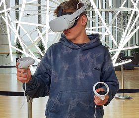 На фото изображен мальчик в очках виртуальной реальности. Он стоит перед футуристически выглядящей лампой.