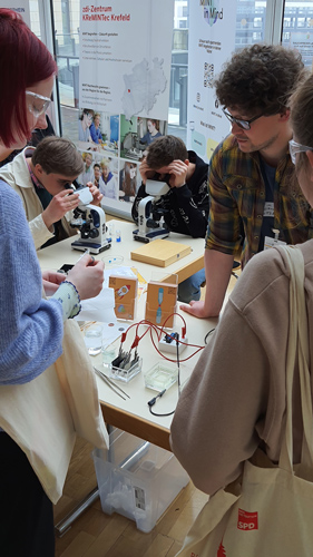 The photo shows a group of young people at a table with scientific experiments. Two boys look into microscopes, two girls look at an experimental setup.