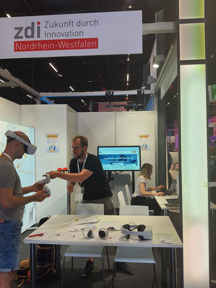 Das Foto zeigt den Stand von zdi.NRW auf der Gamescom. Im Vordergrund erklärt ein Mann einem anderen gerade eine VR-Brille, im Hintergrund sitzt eine Frau an einem Laptop. Auf einem großen Bildschirm steht "Gemeinsam MINT machen".