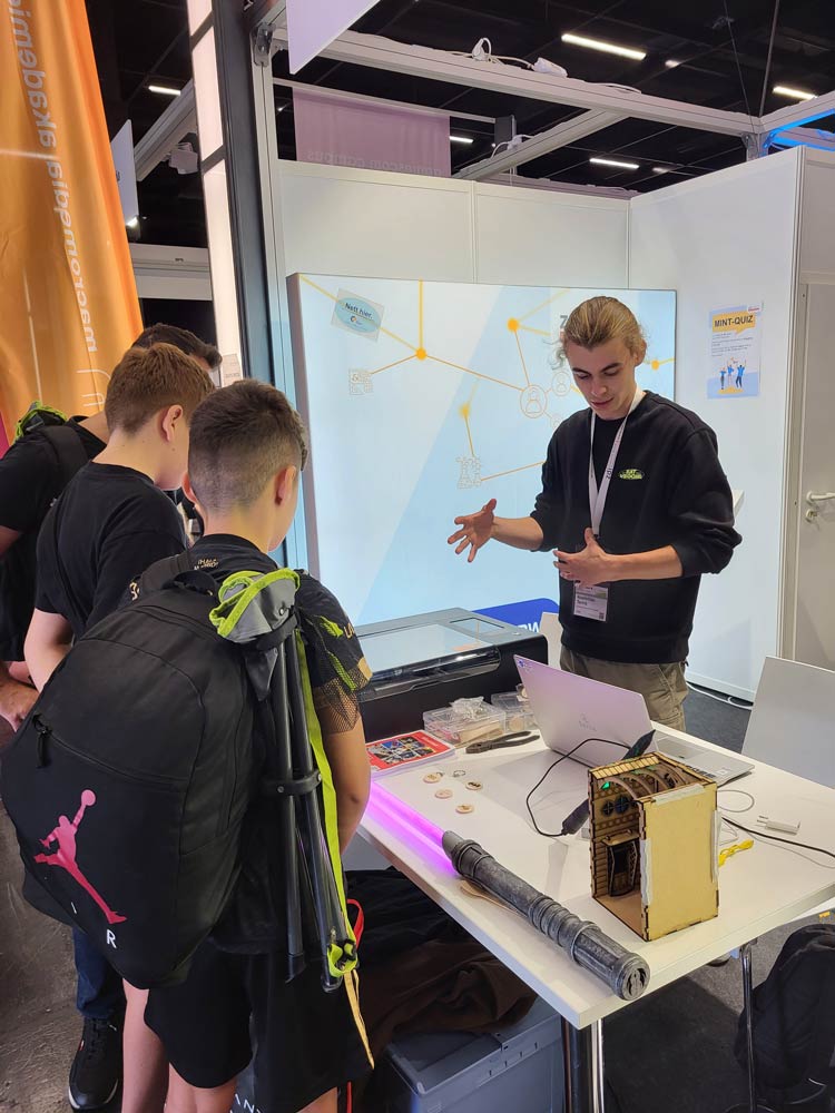The photo shows the zdi.NRW stand at Gamescom. A man is explaining something to three boys, on a table between them are a milling machine and a lightsaber.