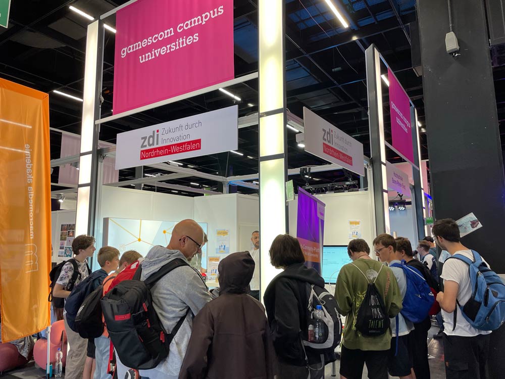 The photo shows the zdi.NRW stand at Gamescom. A sign above the stand reads "gamescom campus universities". There is a crowd of people of different ages around the stand.