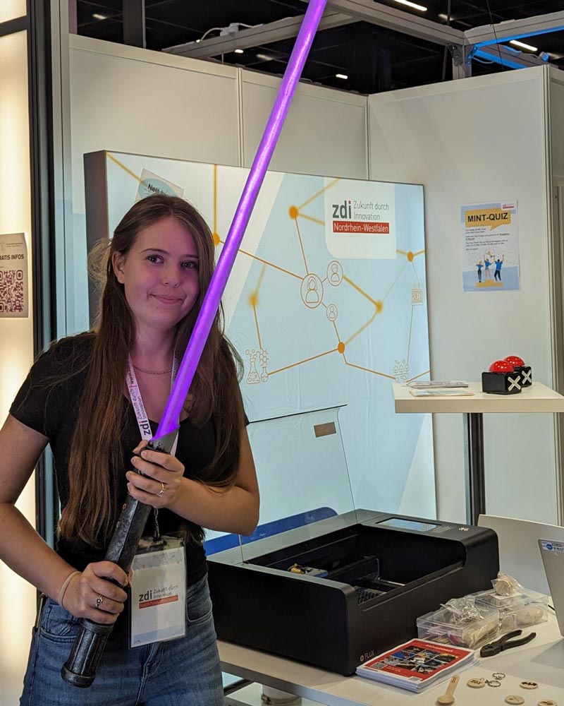 The photo shows Elena from the zdi youth advisory board at the zdi stand at Gamescom. She holds a glowing purple lightsaber in her hands.