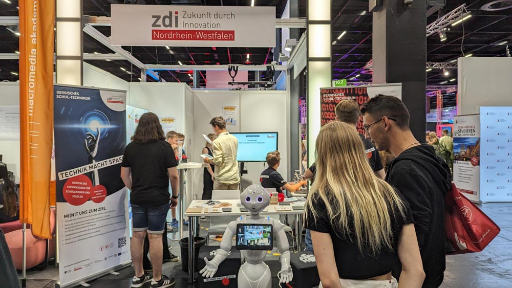 The photo shows the zdi.NRW stand at Gamescom. Several people are standing at the stand and looking at the robot Pepper, which is standing in front of the stand.