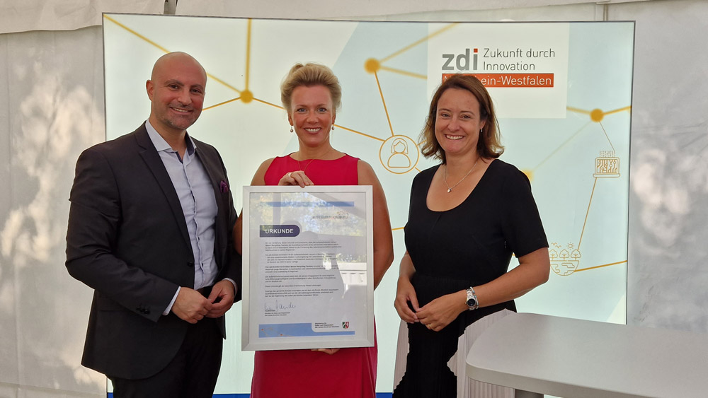 The photo shows District Administrator Ali Doğan, Minister Ina Brandes and District President Anna Katharina Bölling. The minister holds a certificate for the appointment of the zdi student laboratory in her hand. The three are standing in front of a light wall that says zdi - future through innovation North Rhine-Westphalia.
