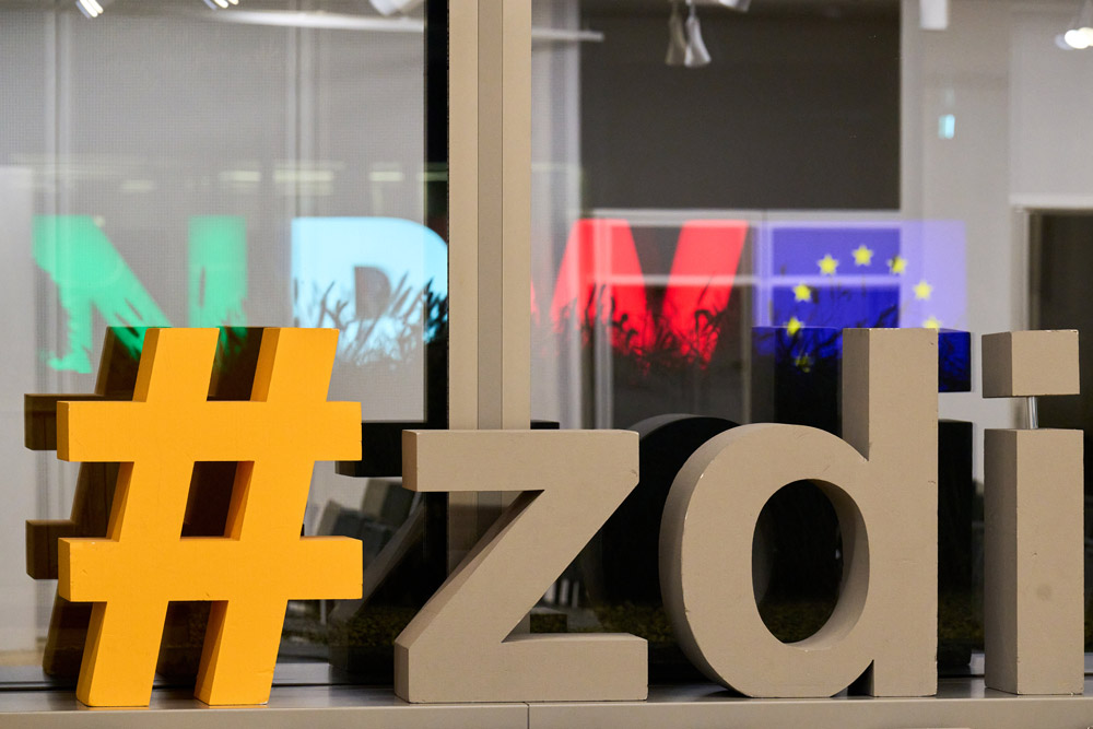 A sign with the inscription “zdi” in front of a window. Through the window you can see the letters NRW in green, white and red and a European flag shining.