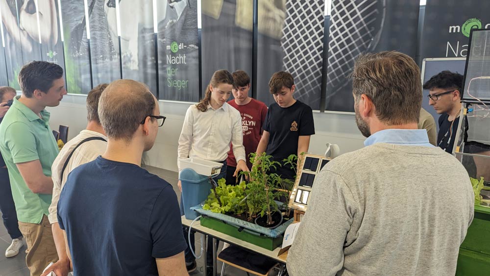 Das Bild zeigt drei Jugendliche, die ihr Science League Projekt der Jury präsentieren. Es ist ein Gewächshaus, in dem einige grüne Pflanzen angebaut sind.