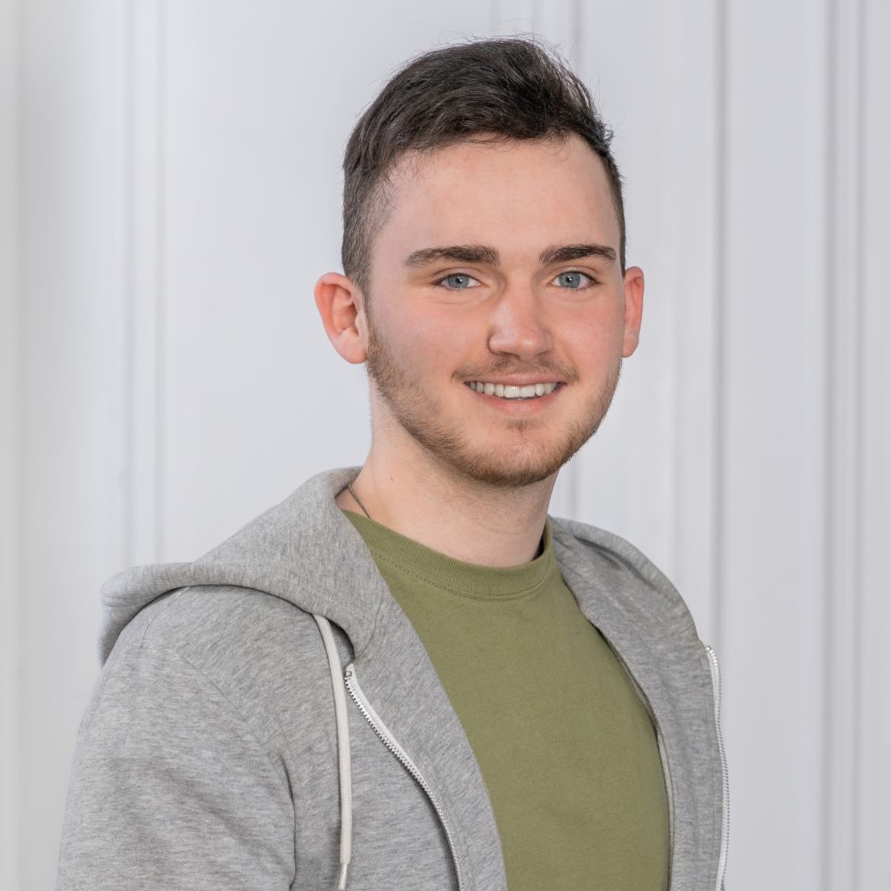 Das Foto zeigt Nicholas Häfner, einen Mann mit kurzen, braunen Haaren und Dreitagebart. Er trägt einen grauen Kapuzenpullover und ein grünes T-Shirt und lächelt freundlich in die Kamera.