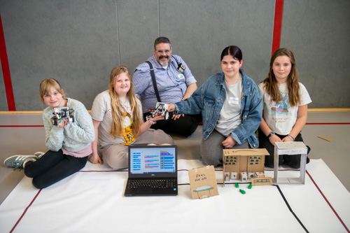 Fünf Personen sitzen im Innenbereich auf dem Boden. Vor ihnen stehen ein Laptop, einige kleine selbstgebaute Modelle und ein Roboter.