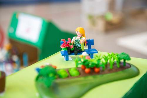 Eine Spielzeugfigur mit blonden Haaren sitzt auf einer blauen Bank und hält rosa Blumen in den Händen. Neben der Figur befindet sich ein kleiner Modell-Gemüsegarten mit grünen Pflanzen und orangefarbenem Gemüse, inspiriert vom zdi-Roboterwettbewerb.