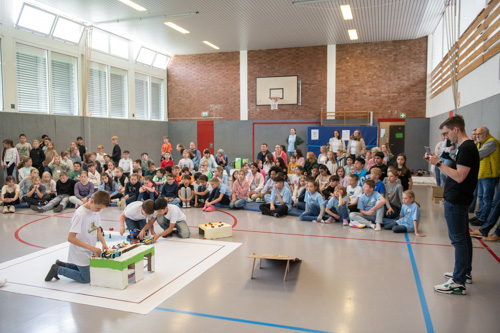Zwei Schülerteams arbeiten im Rahmen des zdi-Roboterwettbewerbs in einer Turnhalle an einem Robotik-Projekt. Eine Gruppe von Kindern und Erwachsenen schaut ihnen zu.