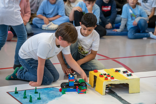 Zwei Jungen knien während des zdi-Roboterwettbewerbs in einer Turnhalle auf dem Boden und bauen LEGO-Modelle zusammen. Im Hintergrund beobachtet eine Gruppe Kinder sie.