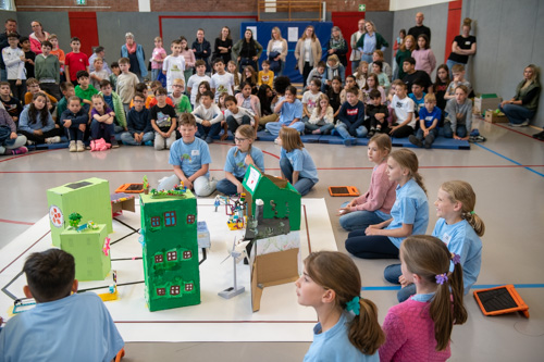 Kinder sitzen in einer Turnhalle im Kreis um grüne Modellbauten. Im Hintergrund beobachtet sie eine große Menschengruppe beim zdi-Roboterwettbewerb.