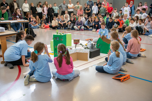 Eine Gruppe von Kindern präsentiert ihr Modellprojekt im Rahmen des zdi-Roboterwettbewerbs auf dem Boden. Ein großes Publikum aus Schülern und Erwachsenen schaut in einer Schulturnhalle zu.