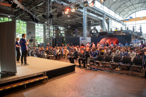 Eine Gruppe von Menschen verfolgt eine Präsentation auf einer Bühne in einem großen Veranstaltungsraum mit Metallträgern und hohen Decken. Der Veranstaltungsort ist im industriellen Stil gehalten.