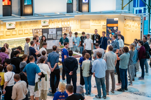 Eine Gruppe von Menschen steht um ein Ausstellungsstück herum, während ein Redner in einem hell erleuchteten Innenraum zu ihnen spricht.