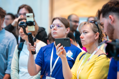Zwei Frauen in einer Menschenmenge filmen oder fotografieren mit ihren Smartphones. Eine Frau nutzt einen Stabilisator. Beide tragen Lanyards und konzentrieren sich auf eine Veranstaltung oder Präsentation.