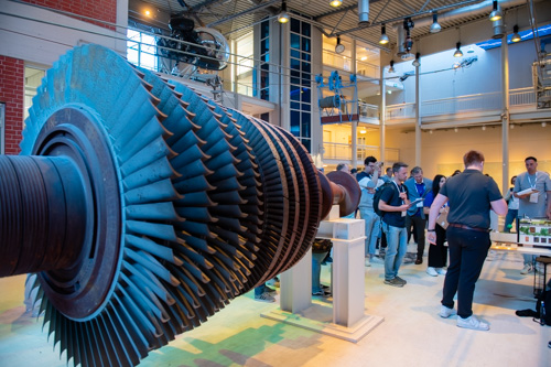 Eine große Industrieturbine wird in einem Innenbereich ausgestellt. Eine Gruppe von Menschen steht in der Nähe und beobachtet und diskutiert in einem gut beleuchteten Ausstellungsraum.