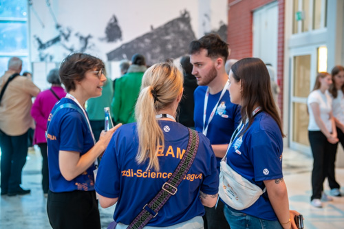Vier Personen in passenden blauen T-Shirts mit der Aufschrift „TEAM“ auf dem Rücken stehen bei einer Veranstaltung zusammen und unterhalten sich. Im Hintergrund sind weitere Personen zu sehen.