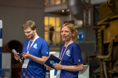 Zwei Personen in blauen Hemden mit Schlüsselbändern stehen drinnen. Eine hält ein Mikrofon und ein Blatt Papier, die andere eine Broschüre. Sie scheinen Gastgeber oder Moderatoren einer Veranstaltung zu sein.