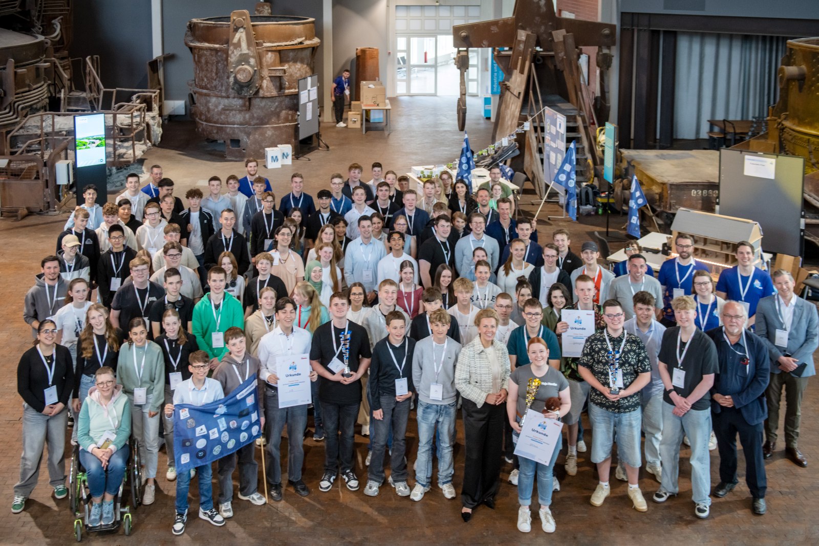 Eine große Gruppe überwiegend junger Erwachsener posiert in einem Industriegebäude für ein Foto. Einige halten Zertifikate und ein Banner in den Händen.