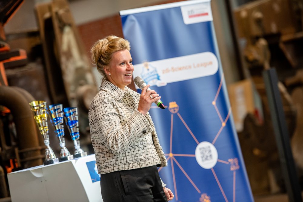Eine Frau spricht bei einer Indoor-Veranstaltung in ein Mikrofon. Auf einem Tisch sind Trophäen ausgestellt, im Hintergrund ist ein blaues Veranstaltungsbanner zu sehen.
