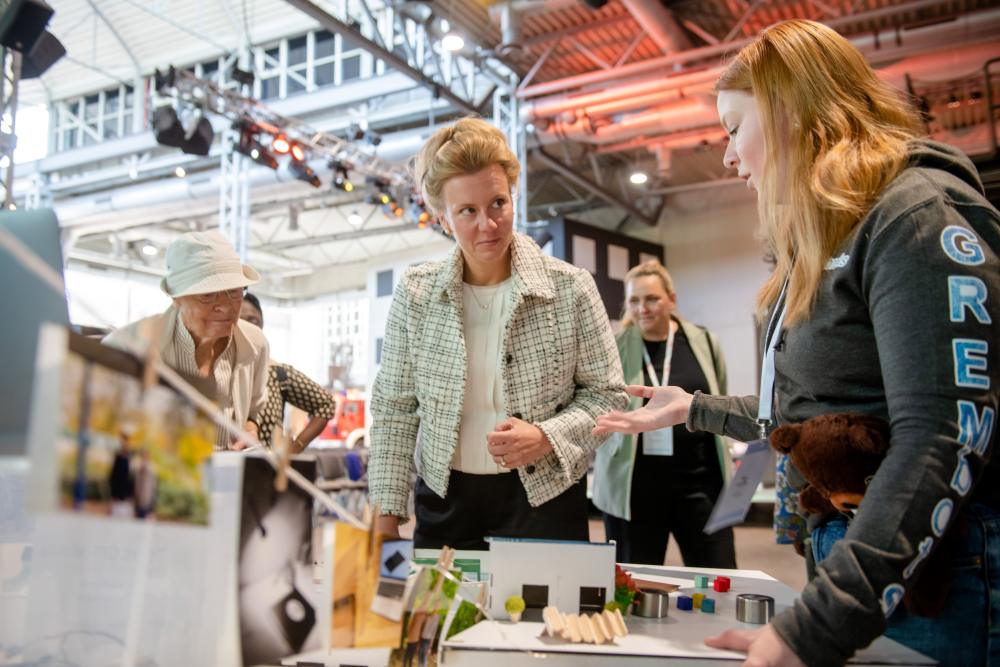 Eine Gruppe von Frauen steht in einem hell erleuchteten Ausstellungsraum um einen Tisch, auf dem Architekturmodelle und Materialien ausgestellt sind.