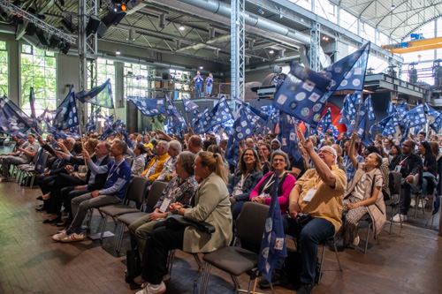 Eine große Gruppe von Menschen sitzt in Stuhlreihen in einem Innenbereich und schwenkt während einer Veranstaltung blaue Fahnen mit weißen Symbolen.