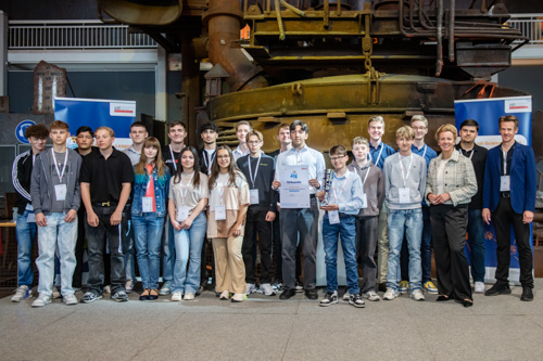 Eine Gruppe Jugendlicher und Erwachsener posiert im Innenbereich für ein Foto. Einige halten Zertifikate in den Händen. Im Hintergrund sind Industrieanlagen zu sehen.
