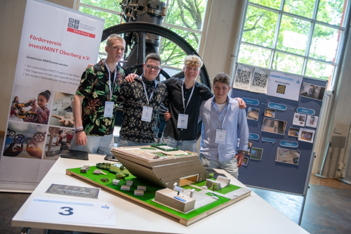 Vier junge Männer stehen hinter einem Architekturmodell. Im Hintergrund sind Plakate und Informationsmaterialien einer Wissenschafts- oder Technikausstellung zu sehen.