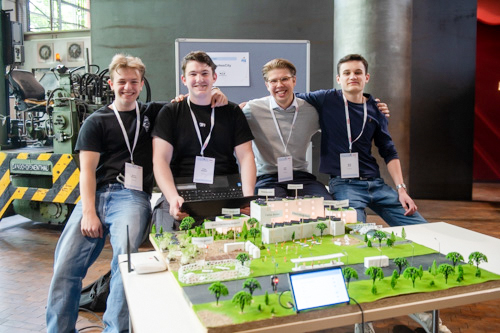 Vier junge Männer mit Namensschildern sitzen und stehen hinter einem detaillierten Architekturmodell eines Gebäudekomplexes. Sie lächeln in die Kamera.