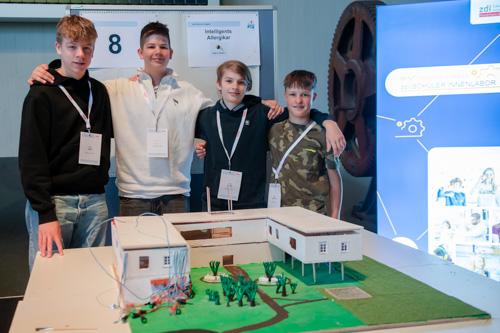 Vier Jungen stehen auf einer Wissenschaftsmesse hinter einem Modellgebäude auf einem Tisch. Sie tragen Namensschilder und lächeln in die Kamera.