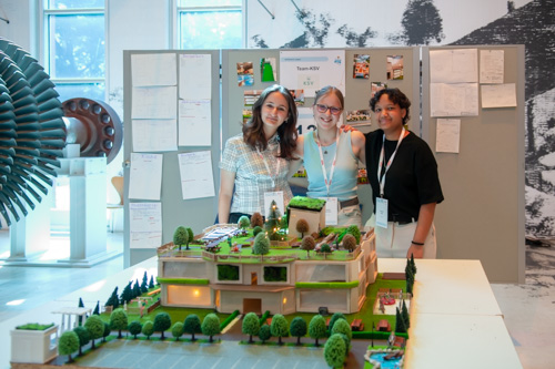 Drei Personen stehen hinter einem detaillierten Architekturmodell eines Gebäudes, das in einer Ausstellungsumgebung auf einem Tisch präsentiert wird.