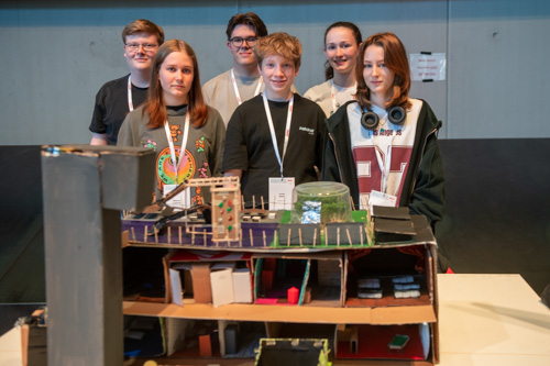 Sechs Teenager stehen drinnen hinter einem Tisch mit einem detaillierten Architekturmodell und posieren für ein Gruppenfoto.