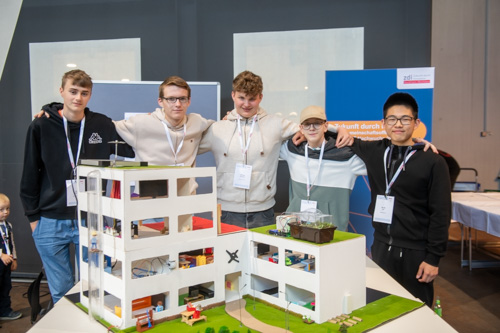 Fünf junge Männer stehen gemeinsam hinter einem Architekturmodell. Sie tragen Namensschilder und Schlüsselbänder. Es scheint sich um eine wissenschaftliche oder technische Veranstaltung zu handeln.