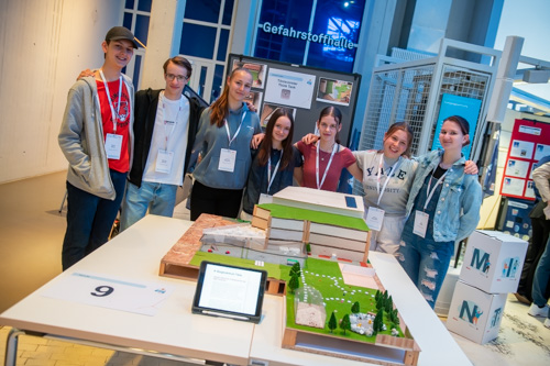 Sieben junge Menschen stehen bei einer Indoor-Veranstaltung hinter einem Tisch, auf dem ein Modellprojekt und Informationsmaterialien präsentiert werden.
