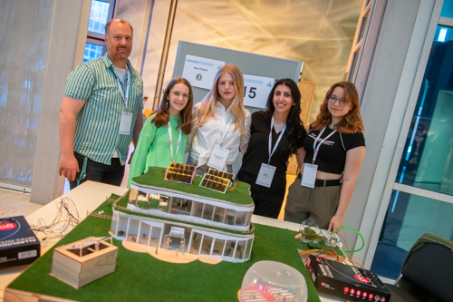 Fünf Personen stehen bei einer Indoor-Veranstaltung hinter einem Tisch, auf dem ein detailliertes Architekturmodell mit Solarmodulen und Verkabelung ausgestellt ist.