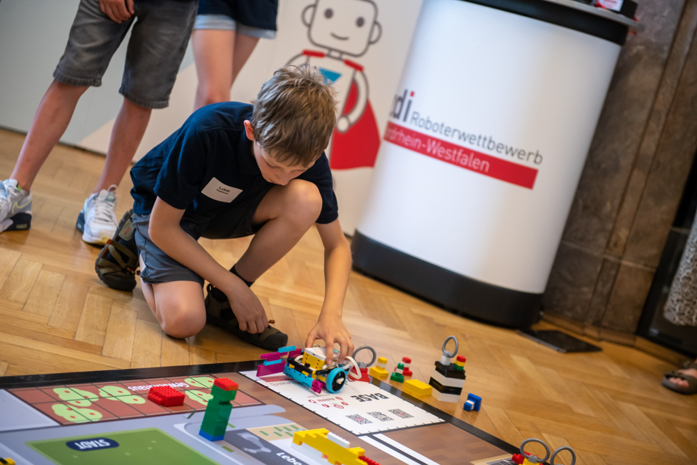 Ein Junge kniet auf dem Boden und justiert einen kleinen Roboter auf einer Wettkampfmatte bei einem Robotik-Event. Im Hintergrund sind weitere Personen und ein Werbebanner zu sehen.