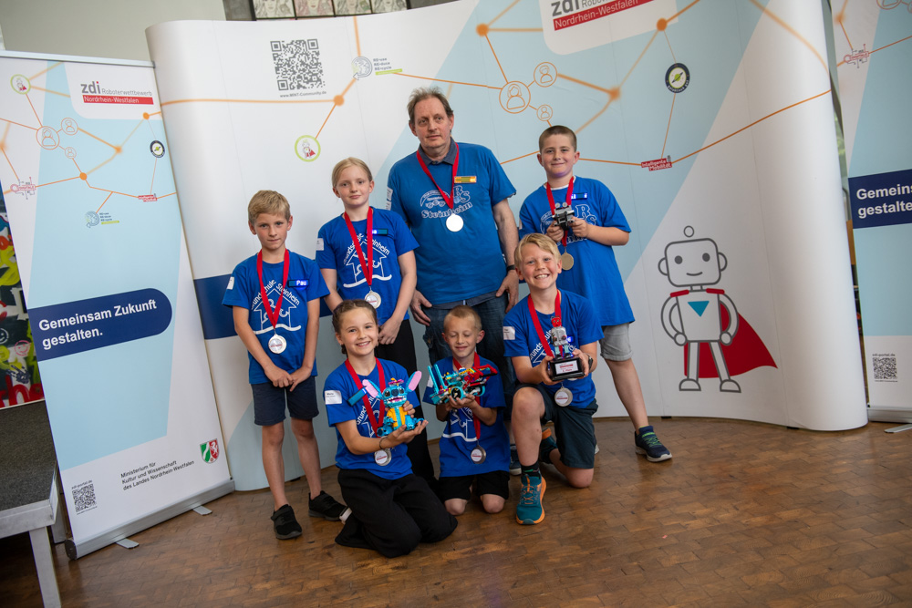 Sieben Kinder und ein Erwachsener, alle in blauen Hemden und Medaillen, stehen mit Trophäen in den Händen im Gebäude. Sie stehen vor Bannern, auf denen wissenschaftliche Grafiken und ein Zeichentrickroboter zu sehen sind.
