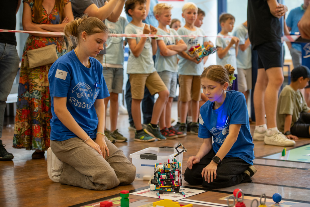 Zwei Mädchen in blauen Shirts basteln bei einem Wettbewerb an einem LEGO-Roboter. Hinter einer Absperrung schaut eine Gruppe von Kindern und Erwachsenen zu.