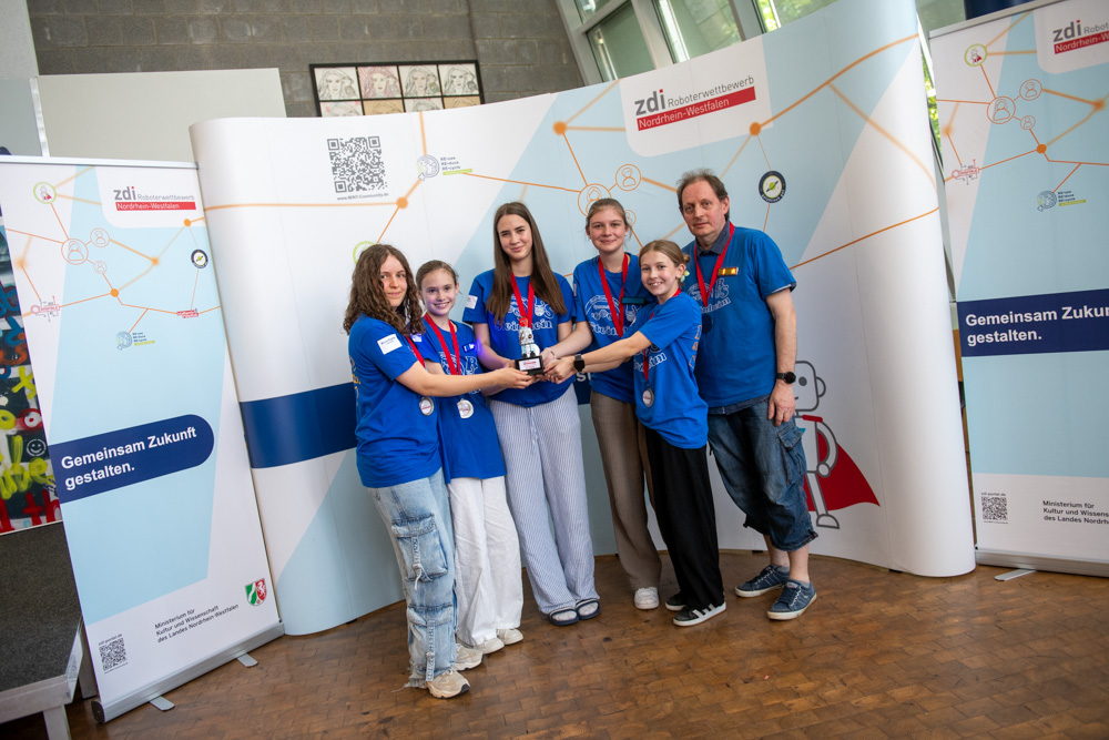 Sechs Personen in blauen Hemden und mit Medaillen stehen bei einer Indoor-Veranstaltung zusammen und halten eine Trophäe vor einem Markenhintergrund.