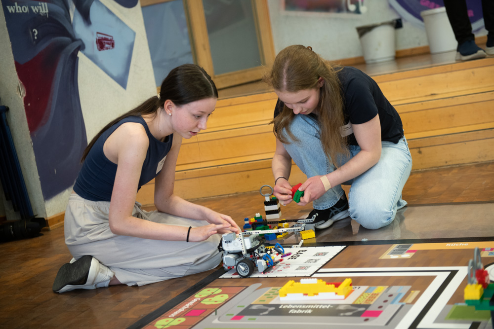 Zwei Mädchen knien in einem Klassenzimmer auf dem Boden. Sie bauen und programmieren auf einer Spielmatte einen LEGO-Robotikbausatz.