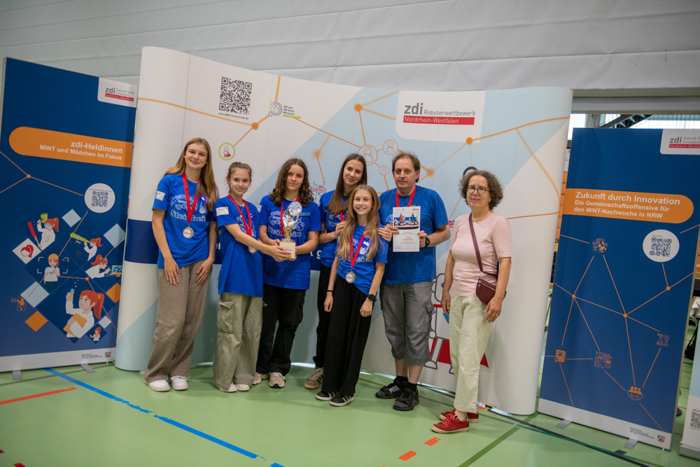 Acht Personen, darunter sechs jugendliche Teilnehmer und zwei Erwachsene, stehen mit Medaillen und einem Pokal in einer Halle. Sie stehen vor Informationsbannern auf einer Wissenschafts- oder Technologieveranstaltung.