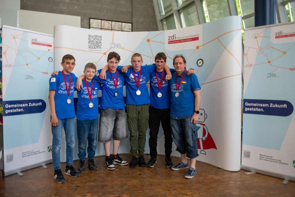 Sechs Jungen und ein Mann tragen passende blaue Shirts und silberne Medaillen. Sie posieren für ein Gruppenfoto vor einem Display mit deutschen Texten und Grafiken.