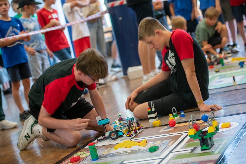 Zwei Jungen knien auf dem Boden und justieren einen Roboter auf einer Wettkampfmatte. Andere Kinder stehen im Hintergrund und schauen bei einem Robotik-Event zu.