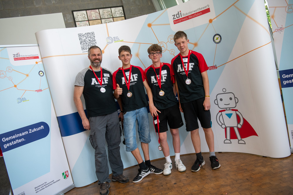 Vier Personen mit Medaillen stehen vor einem Display mit einem Cartoon-Roboter und mehreren Logos. Drei der Personen tragen passende schwarze Shirts mit der Aufschrift „AIF“ auf der Vorderseite.